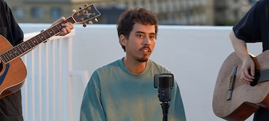 Изображение мужчины, поющего в микрофон на стойке, по обе стороны от мужчины стоят гитаристы