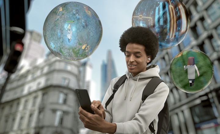 Мужчина с рюкзаком стоит на улице в городе и улыбается, глядя в телефон. Вокруг него парят три пузыря со сценами видеоигры внутри.