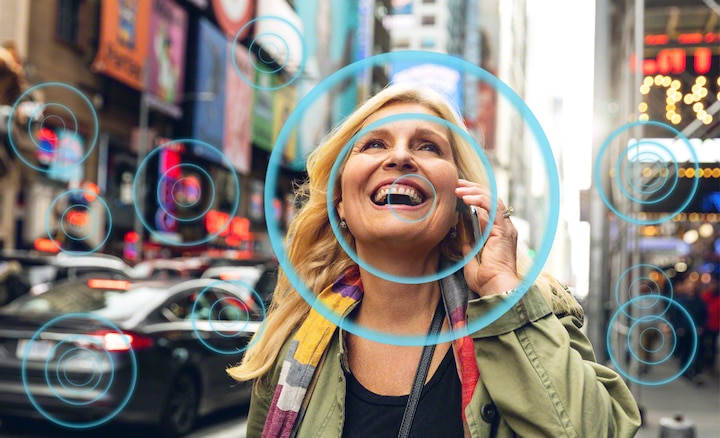 Женщина на оживленной улице, говорящая по телефону, демонстрирует, как технология Voice Zoom 2 улучшает слышимость речи