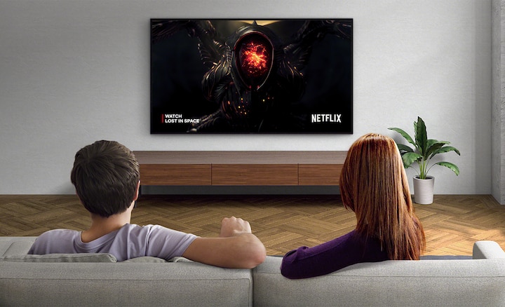 Изображение пары в гостиной, которая смотрит контент Netflix на закрепленном на стене телевизоре BRAVIA. При переключении кнопки Adaptive Calibrated Mode (под изображением) яркость экрана меняется в соответствии с условиями.