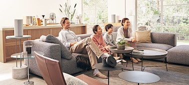 Сидящая на диване семья смотрит телевизор, а динамики, расположенные по всей комнате, создают эффект полного погружения