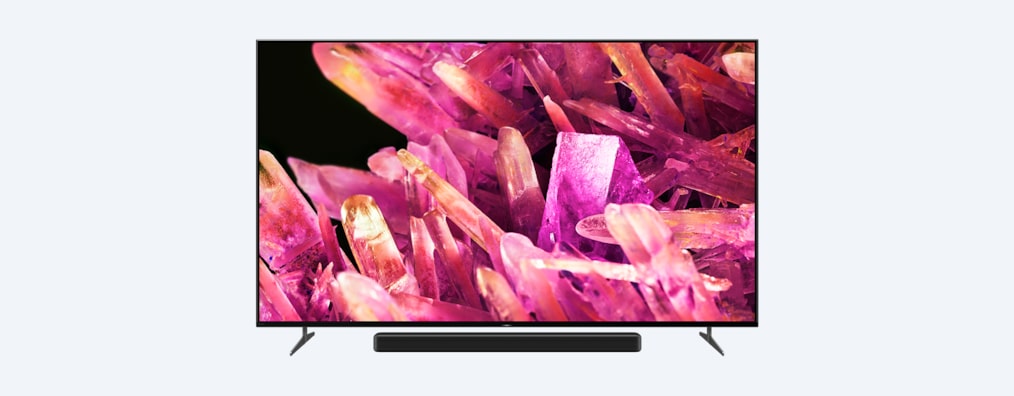 Телевизор BRAVIA X90K на подставке с саундбаром и изображением розовых кристаллов на экране, вид спереди