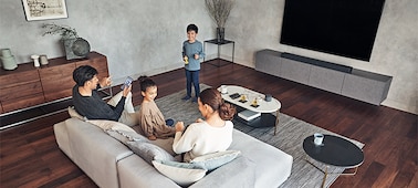 Семья в гостиной наслаждается музыкой. Телевизор и саундбар HT-A7000 установлены на мраморной полке.