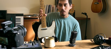 Изображение мужчины, который держит гитару и говорит в находящийся рядом микрофон, при этом мужчина смотрит в камеру, к которой прикреплен приемник