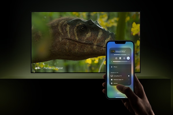 Телевизор на настенном креплении; на экране — голова динозавра; на переднем плане — смартфон в руке с приложением на экране